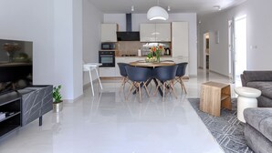Un soggiorno moderno con TV a schermo piatto, una zona pranzo con tavolo e sedie rotonde, una cucina con mobili e elettrodomestici bianchi, e un divano grigio.