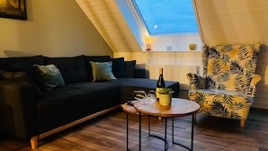 A cozy living room with a dark sofa, a round wooden table with a bottle and glasses, a patterned armchair, and a skylight. - 4-guest Elegant Holiday Base in Hotton (Hotton)