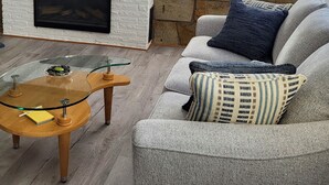 Un salón con pared de piedra, chimenea, una mesa de centro de cristal y un sofá gris con cojines.