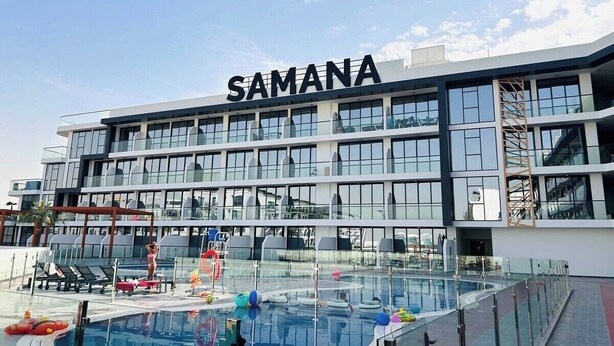Ein modernes Gebäude mit der Aufschrift 'SAMANA' auf der Fassade, ein Schwimmbecken mit Liegestühlen und einem klaren Himmel.