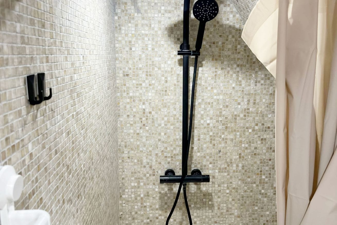 Ducha con manguera negra y rociador redondo, paredes de azulejos y un perchero blanco.
