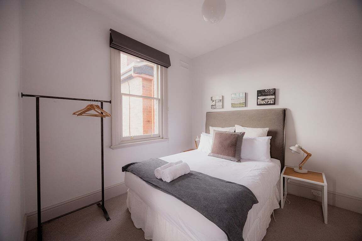 A bedroom with a bed, a bedside table, a hanging clothes rack, and a window with blinds.