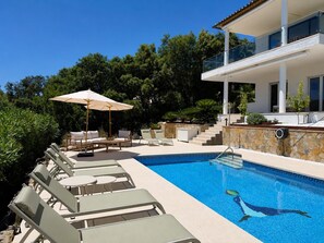 A swimming pool with lounge chairs and an umbrella, surrounded by greenery and a modern building. - Villa Belleza - Santa Cristina d'Aro, Catalonia (Santa Cristina d'Aro)