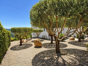 A landscaped garden with neatly trimmed hedges, a large rock centerpiece, and a white building with a visible window. - Villa Belleza - Santa Cristina d'Aro, Catalonia (Santa Cristina d'Aro)