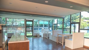 Amplia habitación con un mostrador de recepción de madera, asientos blancos, ventanas grandes y vistas al exterior.
