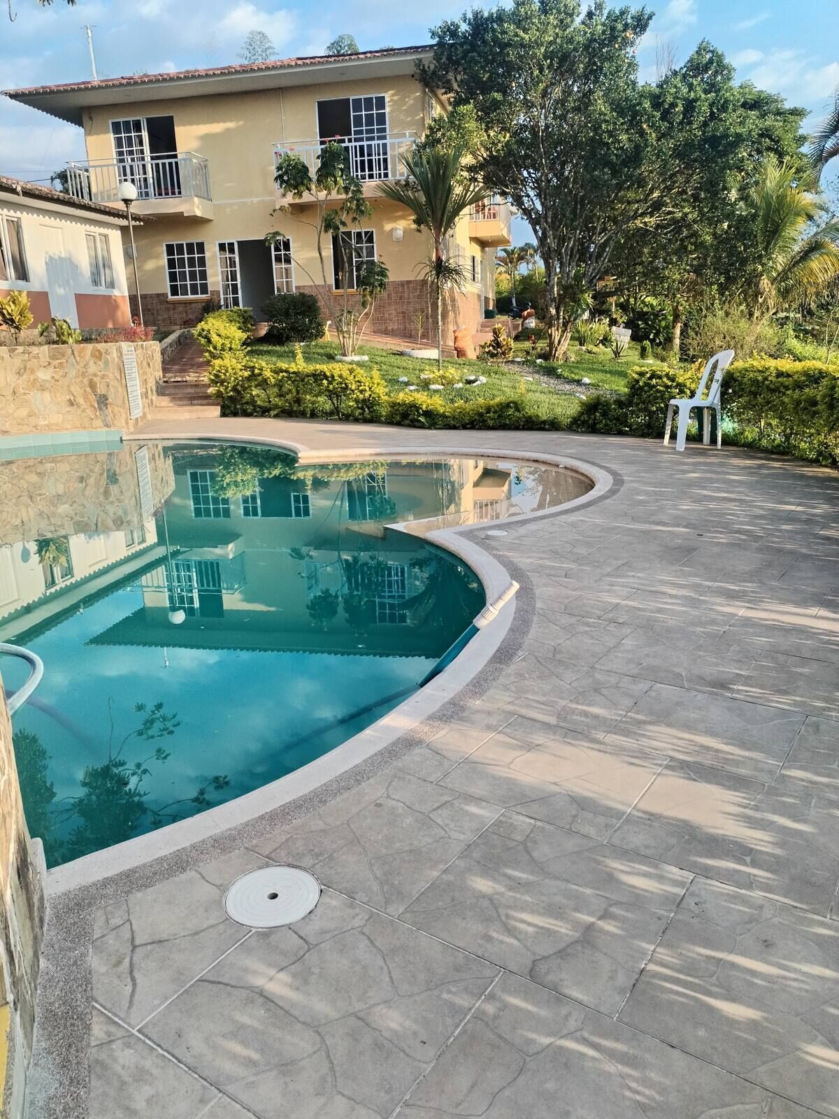 Una piscina con bordo curvo, un patio in pietra e una casa sullo sfondo con un balcone.