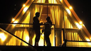 Ein dreieckiges Gebäude mit einem großen Fenster und Vorhängen, beleuchtet von warmem Licht.