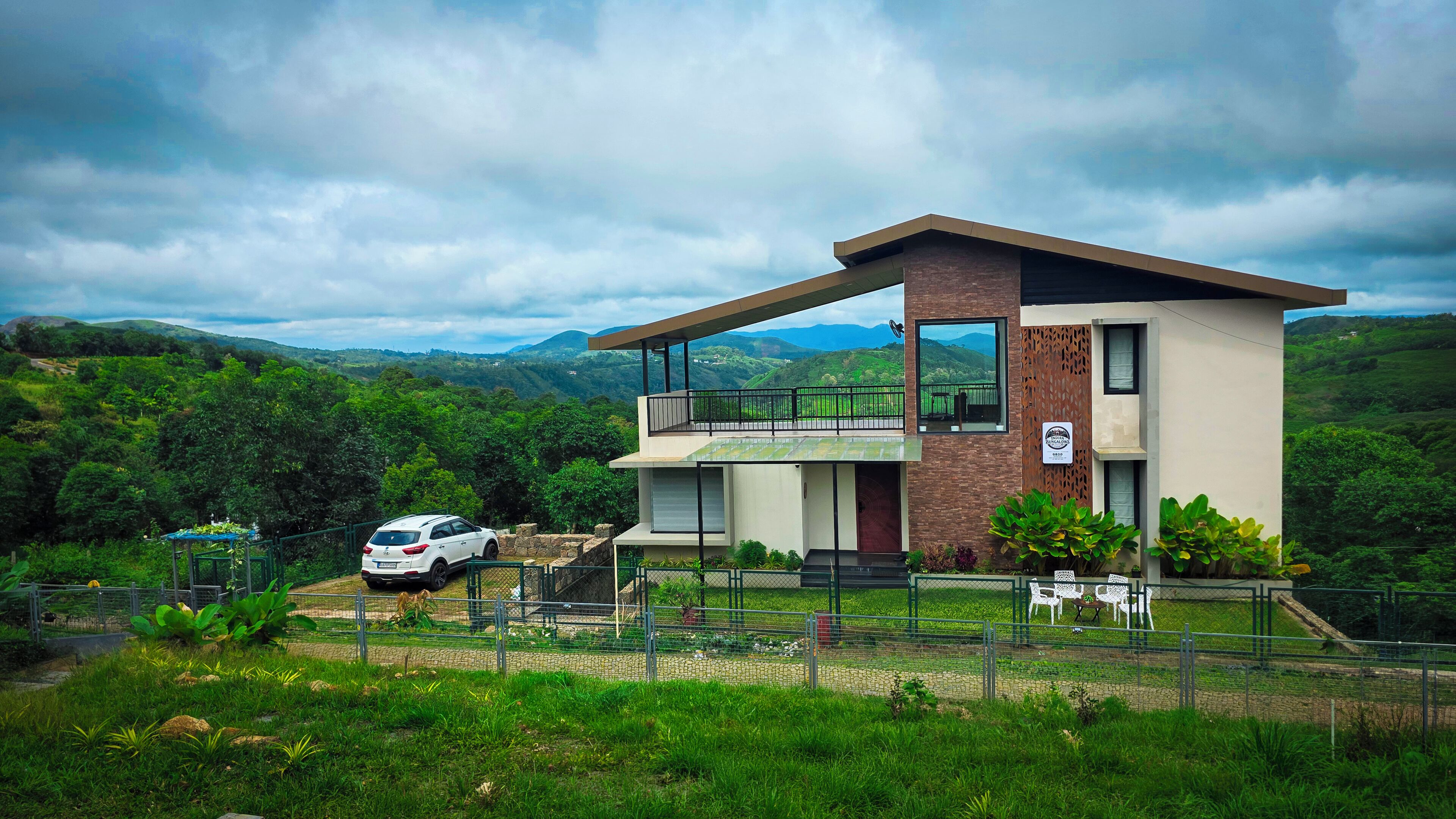 Um edifício moderno de dois andares com varanda, cercado por vegetação exuberante e montanhas ao fundo.