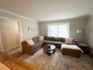 A living room with a sectional sofa, a round coffee table, and a large window with blinds. - Minutes to Arrowhead! 4BR Home with pool table, finished basement & Fire Pit (Kansas City)
