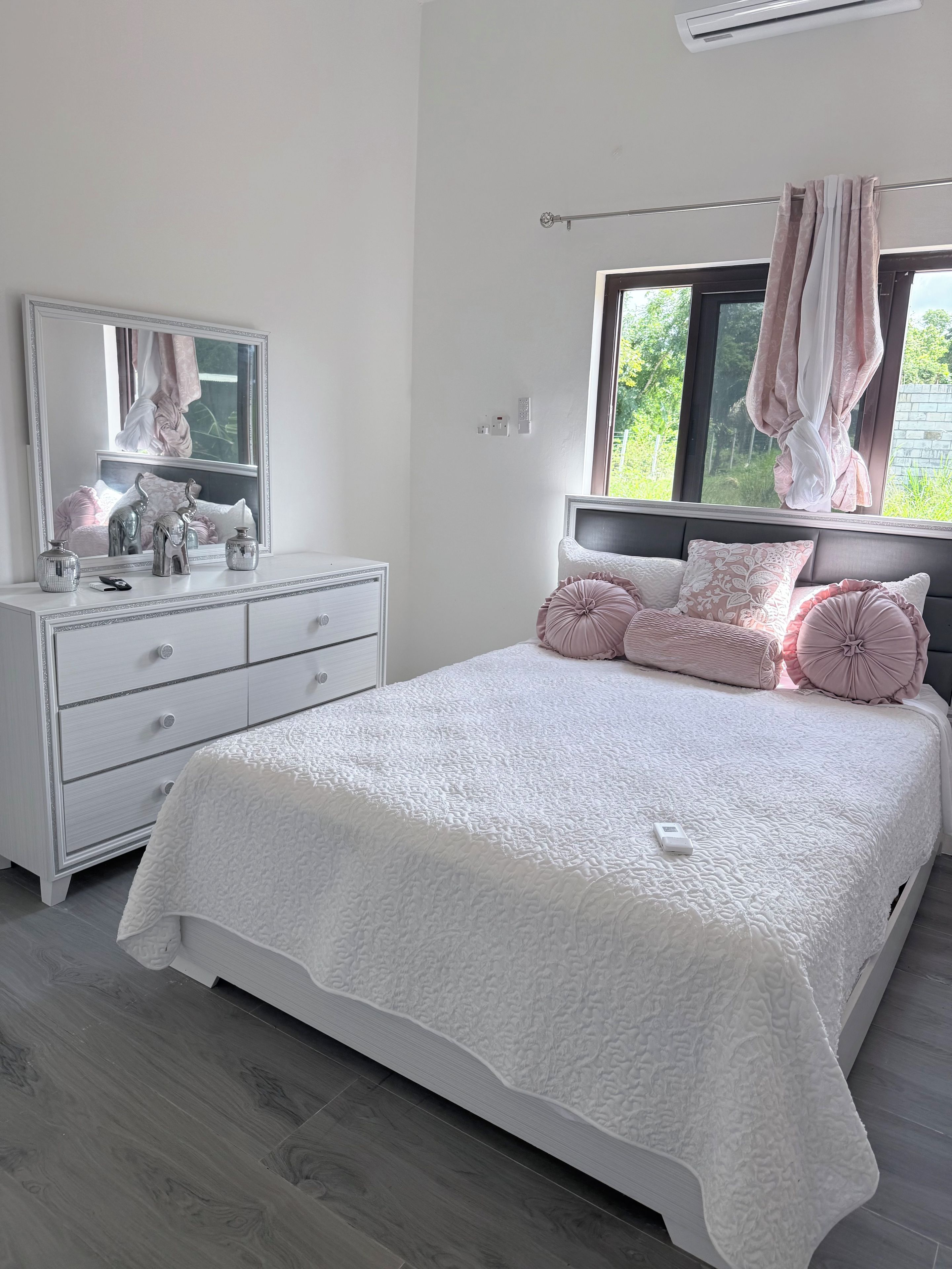 Ein Schlafzimmer mit einem weißen Bett, eine Kommode mit Schubladen, einem Spiegel und einem Fenster mit Vorhängen.