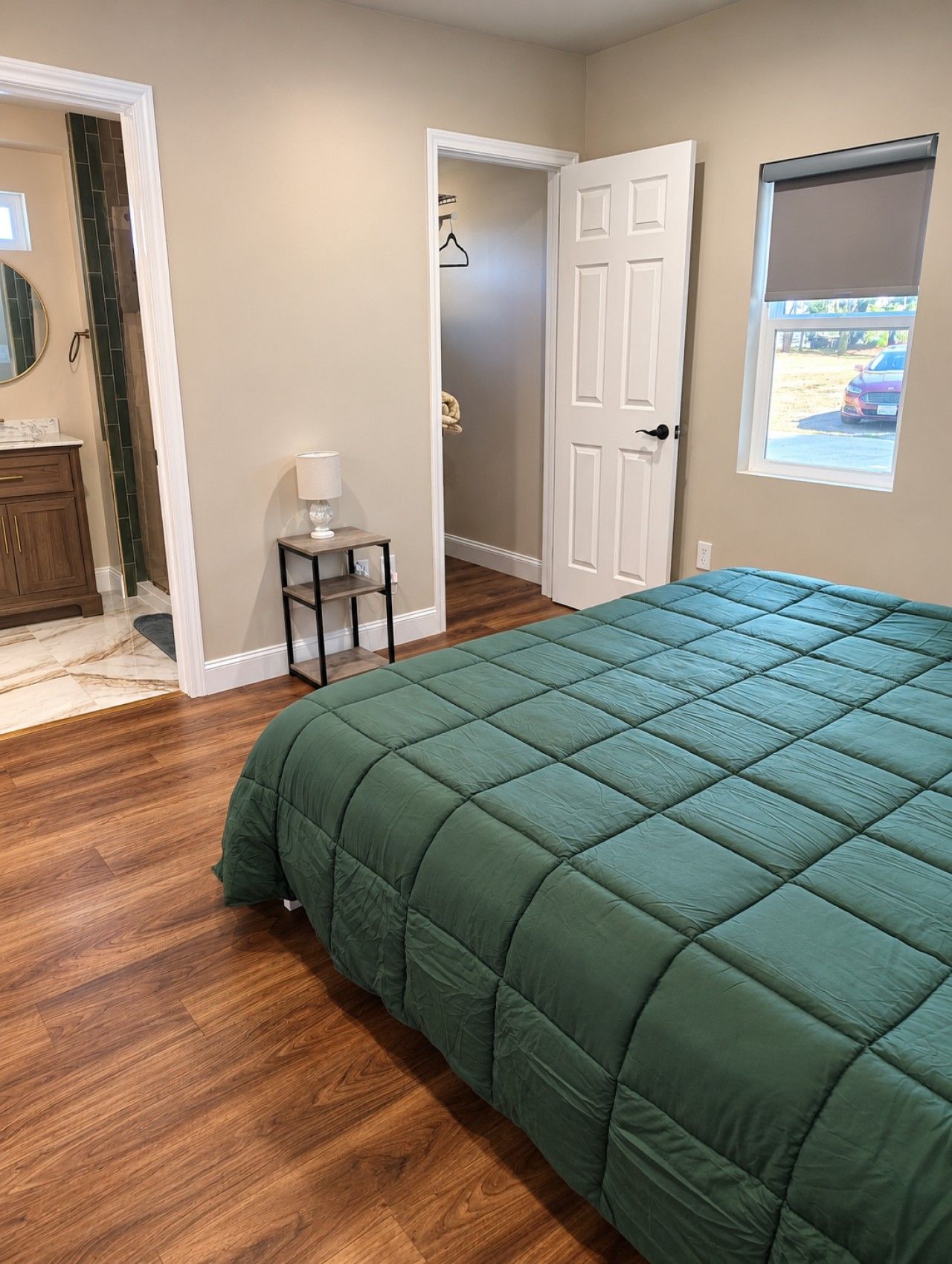 Une chambre avec un couvre-lit matelassé vert, un plancher en bois, une petite table de chevet avec une lampe, une porte donnant sur une autre pièce et une fenêtre avec vue sur une voiture garée.