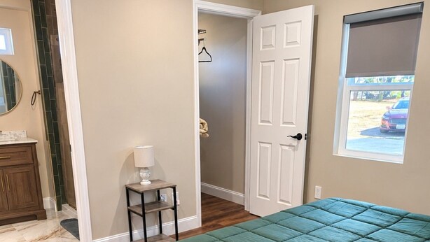 Quarto com colcha verde acolchoada, piso de madeira, mesa de cabeceira pequena com luminária, porta que dá para outro cômodo e janela com vista para um carro estacionado.
