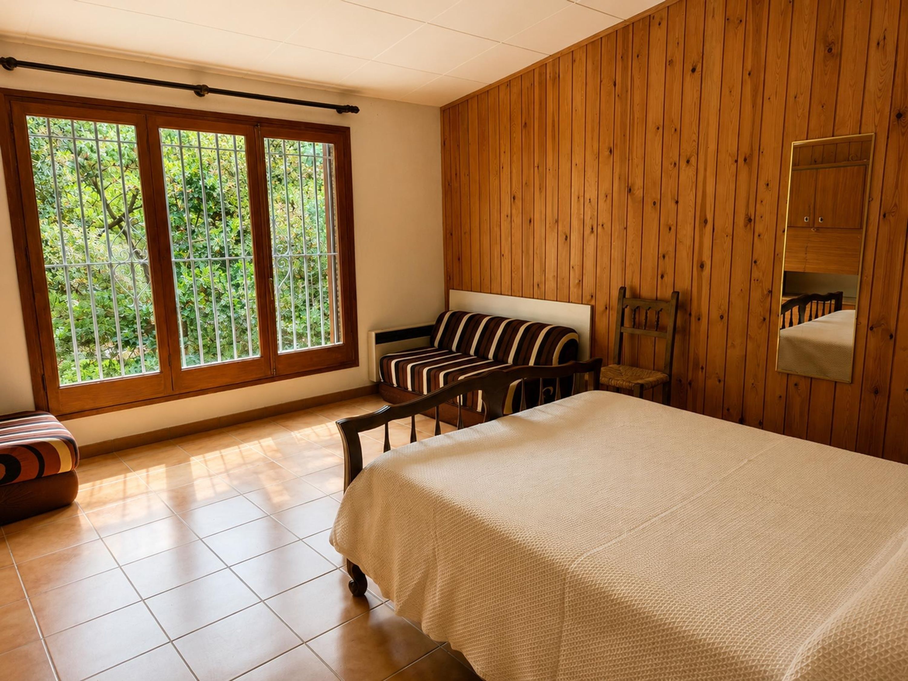 Un dormitorio con cama, una silla, un banco, pared de madera y ventana con vistas a zonas verdes.
