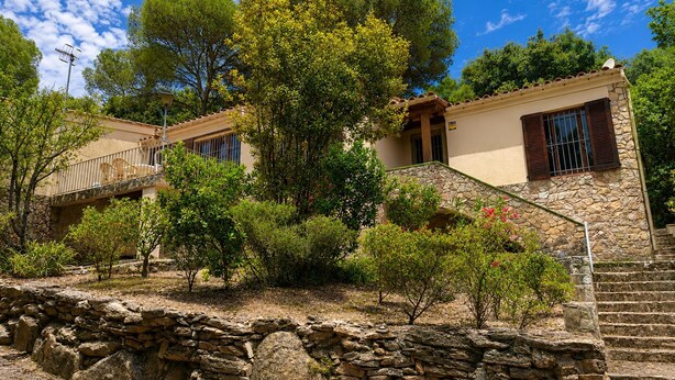 A stone-walled building with a balcony, a staircase, and a tree in front.
