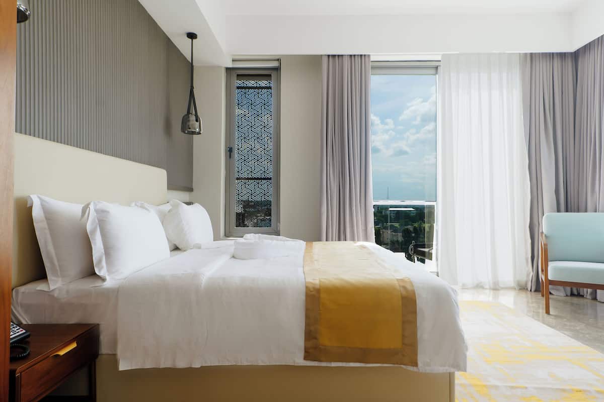 Habitación de hotel con cama, escritorio, silla, ventana con cortinas y vista a la ciudad.