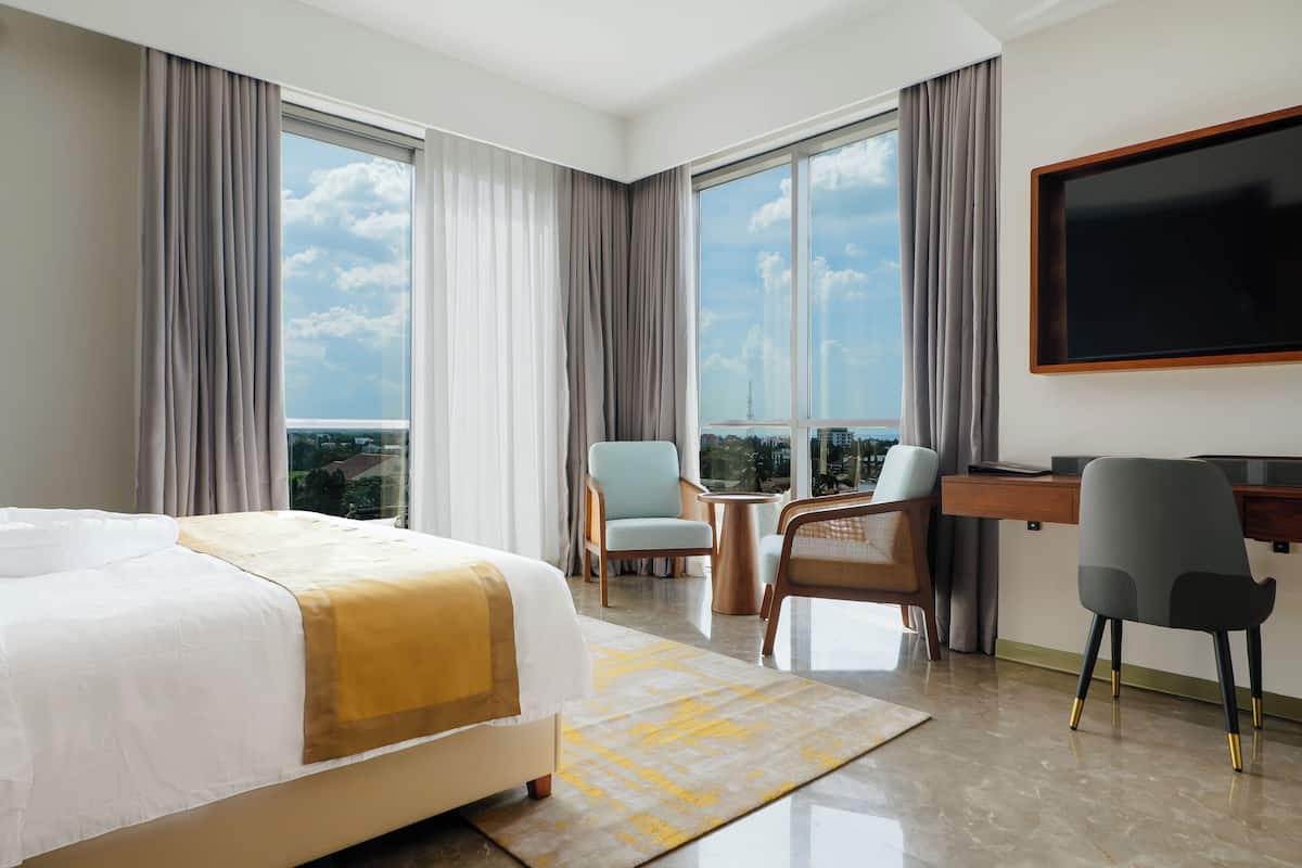 Una habitación de hotel con cama, escritorio, dos sillas, televisor y ventanas amplias.