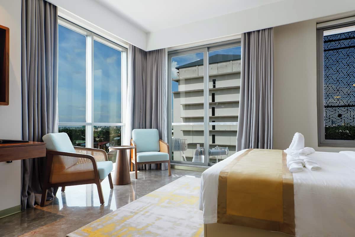 Una habitación de hotel con una cama, dos sillones, un escritorio y ventanas grandes.