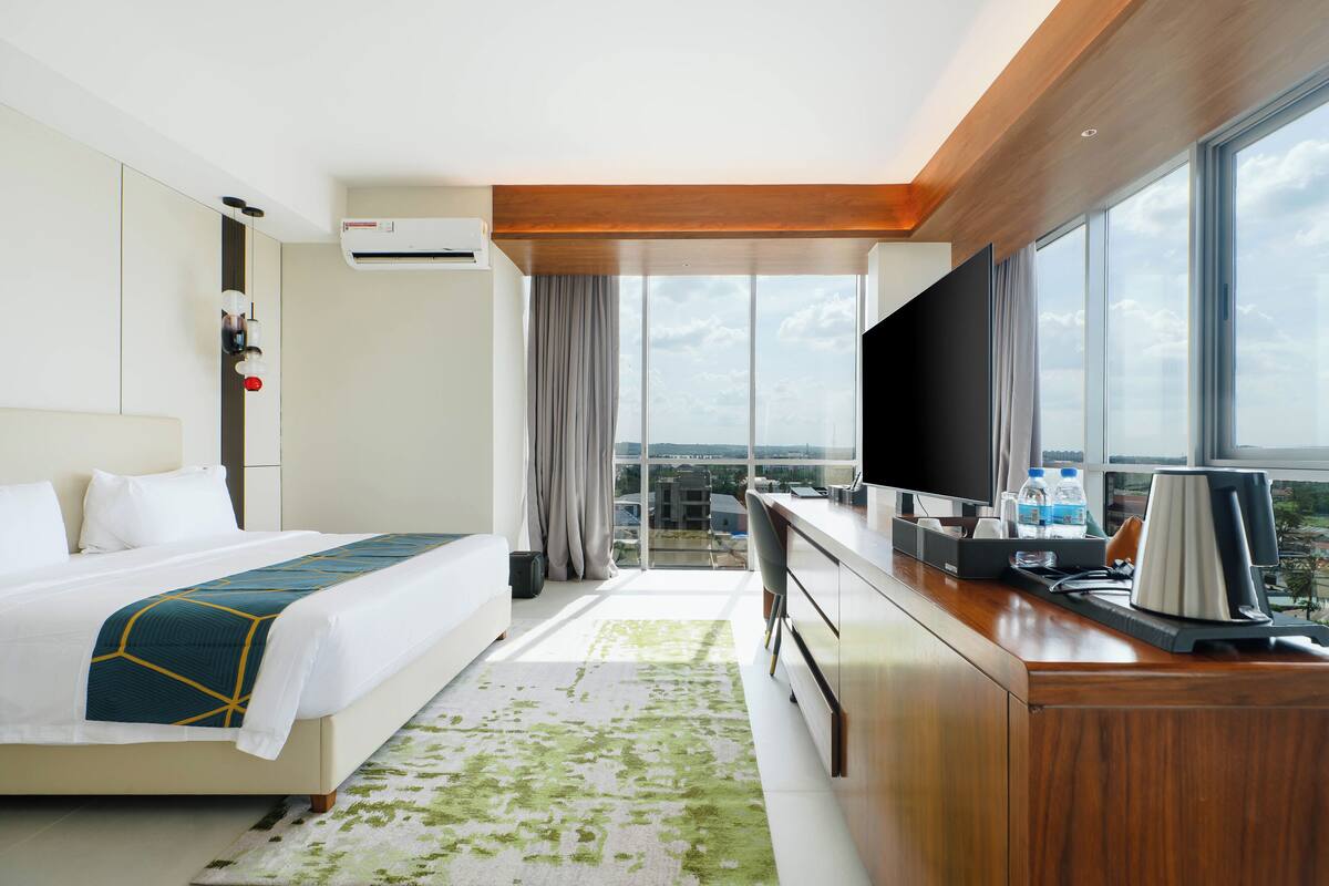 Habitación de hotel con cama, escritorio con computadora, televisor y ventana grande con vista a la ciudad.