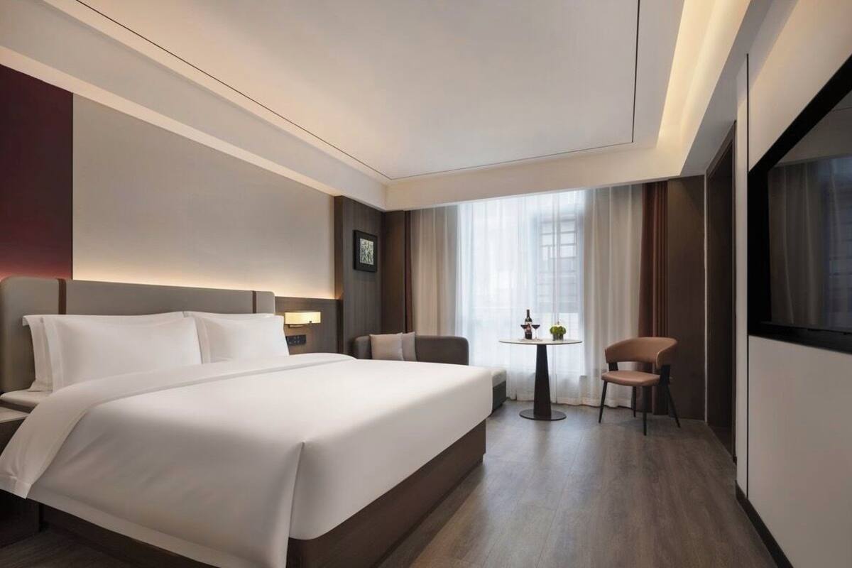 Habitación de hotel con una cama grande, una mesita pequeña con dos sillas, un televisor y una ventana con cortinas.