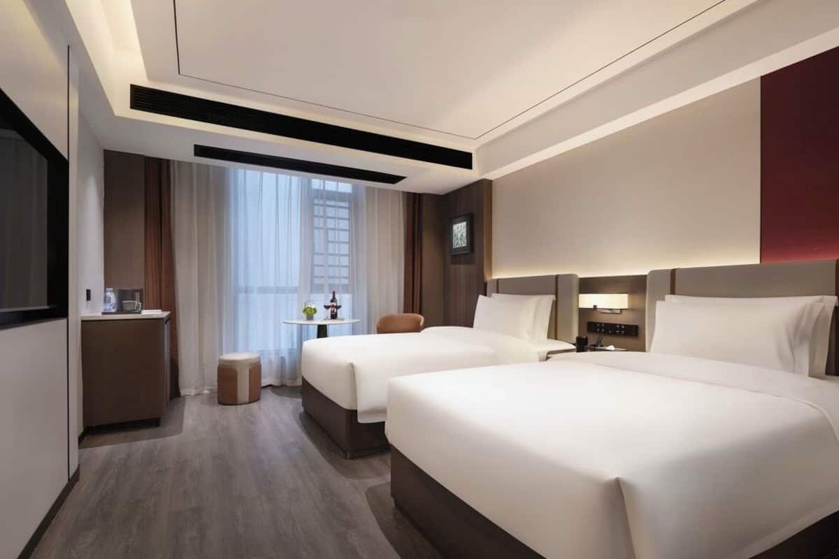 Habitación de hotel con dos camas, una mesita con una lámpara, una ventana con cortinas y un televisor montado en la pared.
