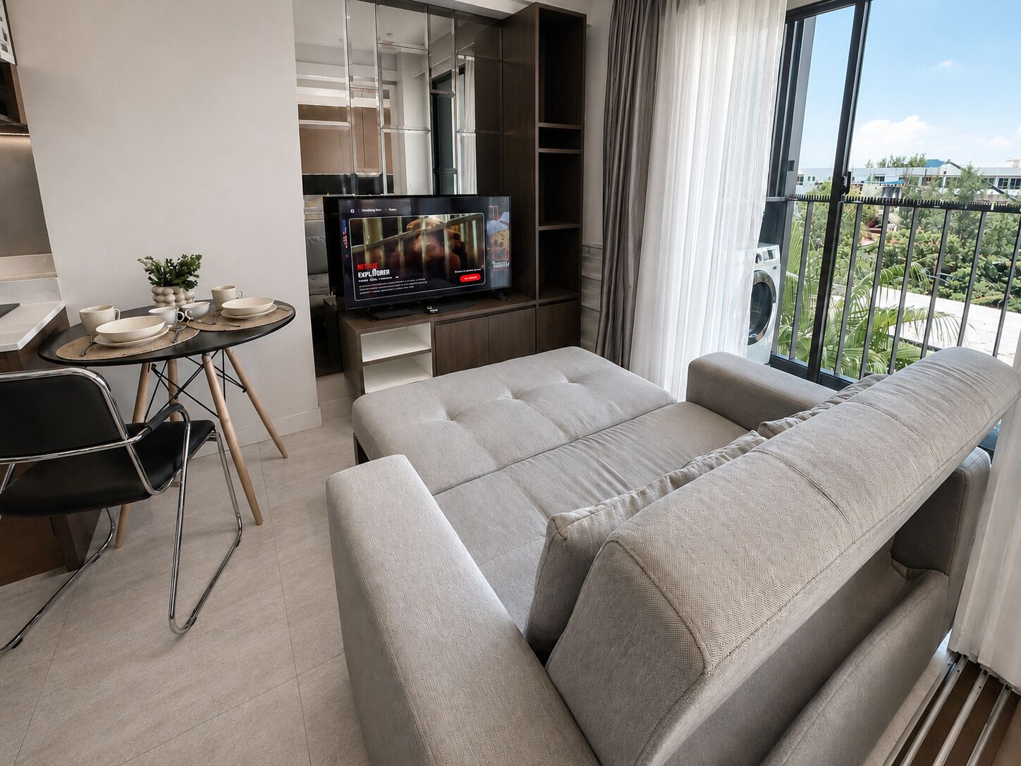 A modern living room with a sofa, a dining area with a table and chairs, a TV, and a balcony with a view.
