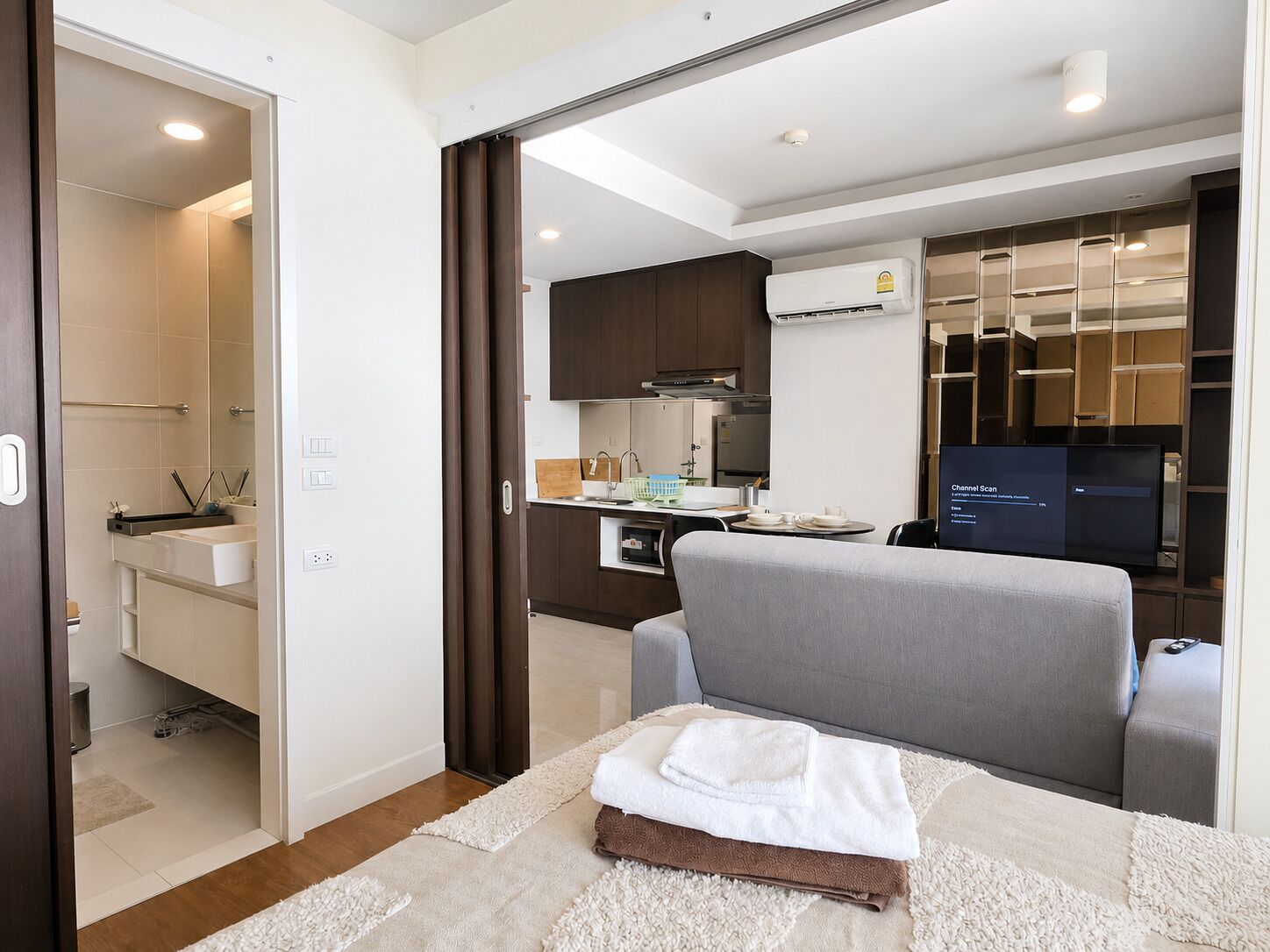 A modern hotel room with a bed, a grey sofa, a small kitchen area, and a bathroom.