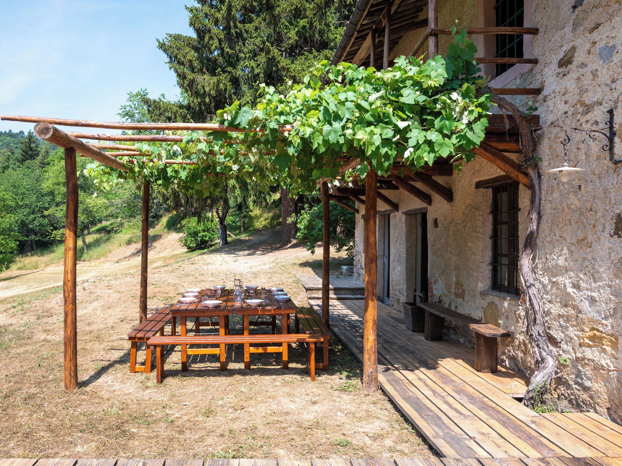 Un área de comedor al aire libre de estilo rústico, con un porche, una mesa de madera y bancos, frente a un edificio de piedra con vistas a una ladera boscosa.