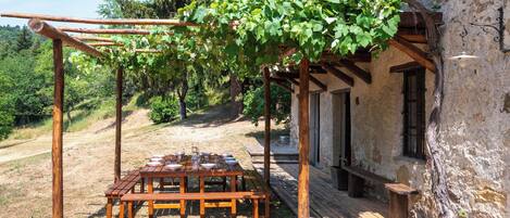 Un área de comedor al aire libre de estilo rústico, con un porche, una mesa de madera y bancos, frente a un edificio de piedra con vistas a una ladera boscosa.