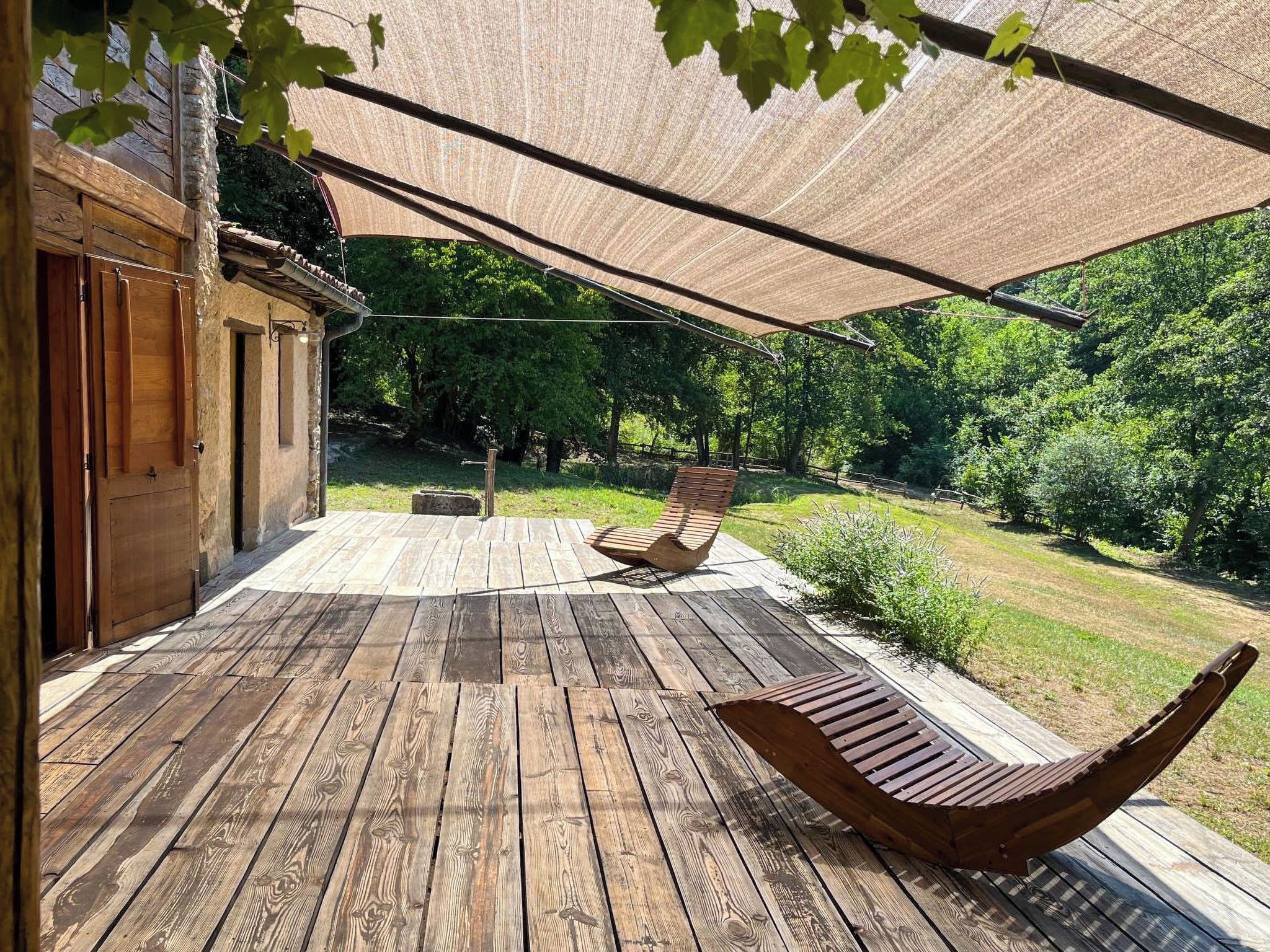 Une terrasse en bois avec un hamac et un banc, entourée de verdure et d’un bâtiment.