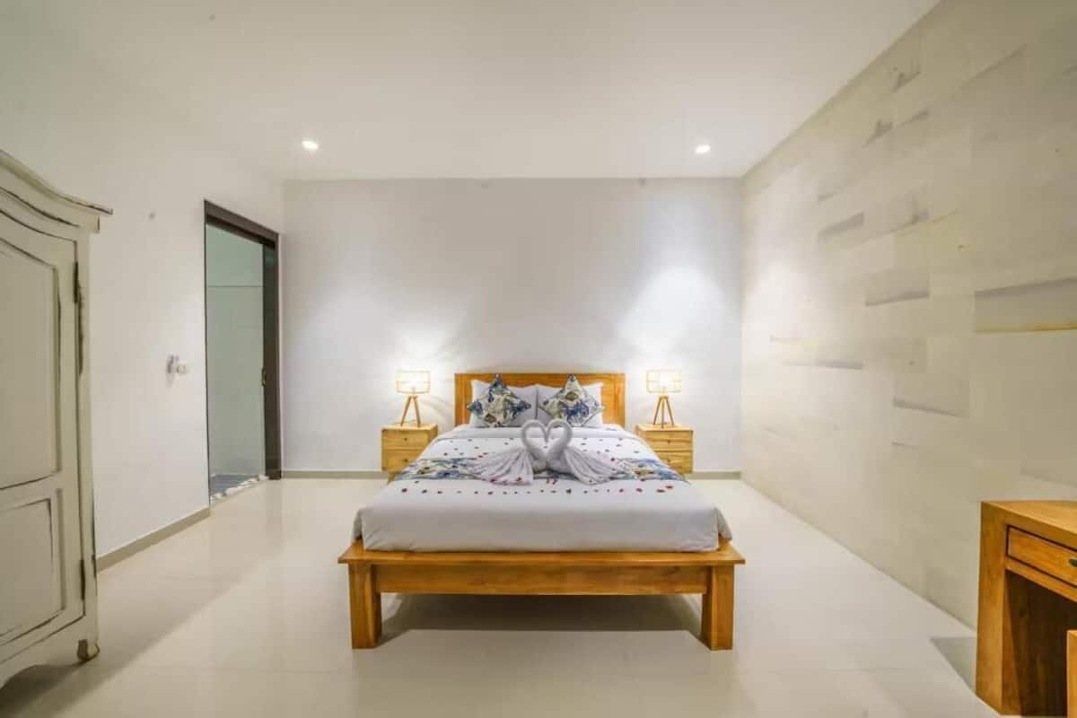 Un dormitorio con una cama de madera, dos lámparas de noche, una toalla decorativa con forma de cisne y una pared lisa.
