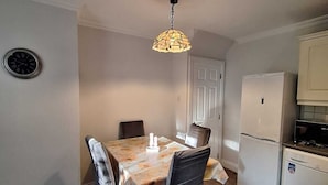 A dining area with a table, chairs, a hanging light fixture, a clock, and a kitchen with a refrigerator.