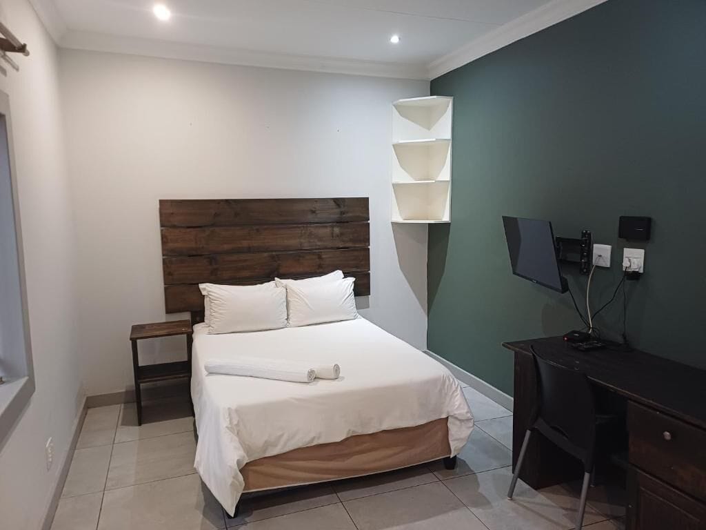 A bedroom with a wooden headboard, a bed with white linens, a desk with a computer, and a wall-mounted TV.