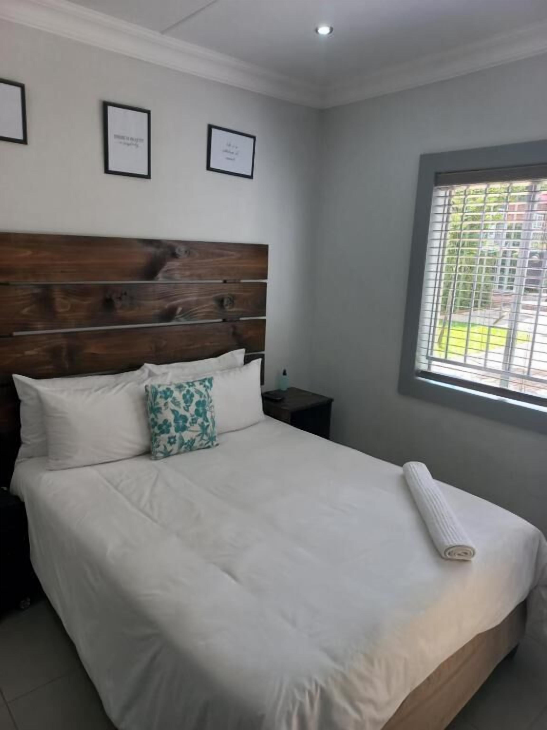 A bedroom with a wooden headboard, white bedding, a teal pillow, a rolled towel, a nightstand, and a window with a view.