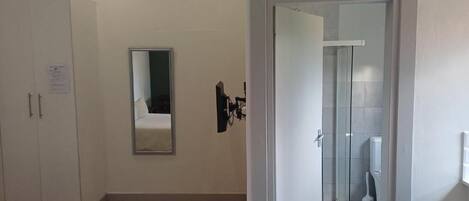 A hotel room with a bed, a wardrobe, a mirror, and a door leading to a bathroom with a shower and toilet.