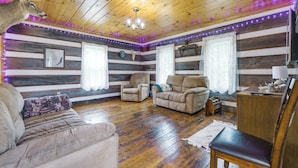 Ein Zimmer mit Holzwänden, Holzboden, einer Couch, zwei Sesseln, einem kleinen Tisch und einem Fernseher.