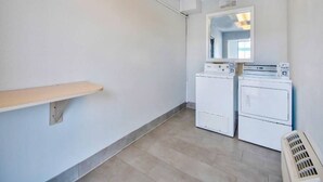 A laundry room with two stacked washers and dryers, a mirror above the dryers, and a wooden shelf on the left wall.