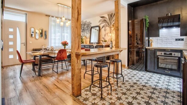 A modern kitchen with a wooden bar, bar stools, and a dining area with a red chair. - A Getaway in the Cotentin (Cherbourg-en-Cotentin)