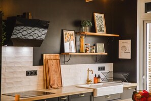 A modern kitchen with a white tiled backsplash, a wooden countertop, and a built-in stove. There are shelves above the sink holding various kitchen items and framed artwork. - A Getaway in the Cotentin (Cherbourg-en-Cotentin)