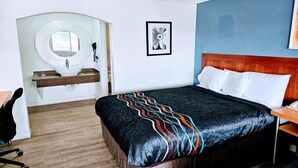 A hotel room with a bed, a round mirror, a sink, a framed picture, and a wooden headboard. - King Bed|Non-Smoking (Ridgecrest CA)