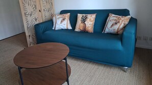 Ein blaugrünes Sofa mit drei gemusterten Kissen, ein kleiner runder Holztisch und eine dekorative Holzvertäfelung mit Blattmotiven.