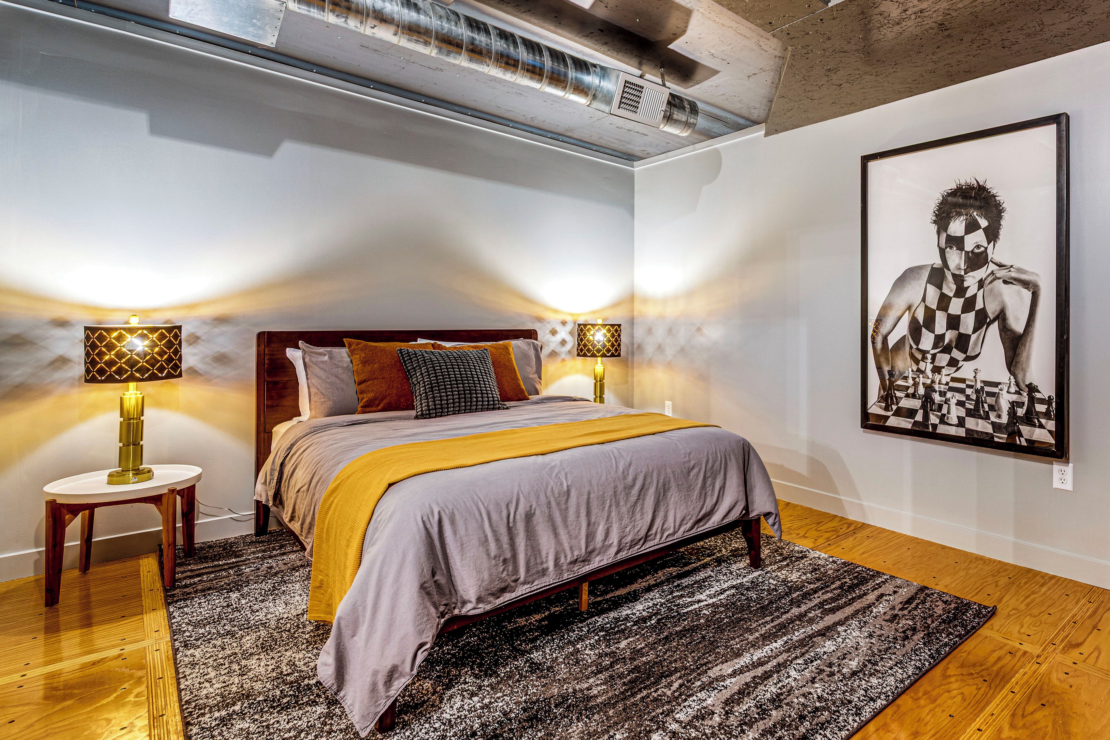 A bedroom with a bed, two bedside lamps, a checkered artwork on the wall, and a patterned rug.