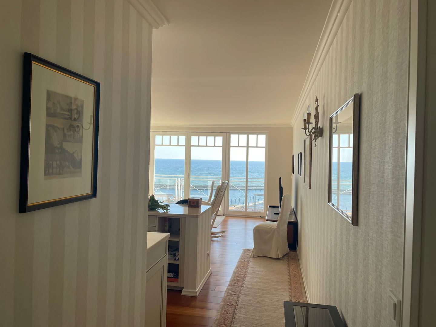 Ein Flur mit Blick auf das Meer, gerahmten Kunstwerken und einer Sitzecke mit einem Sessel.
