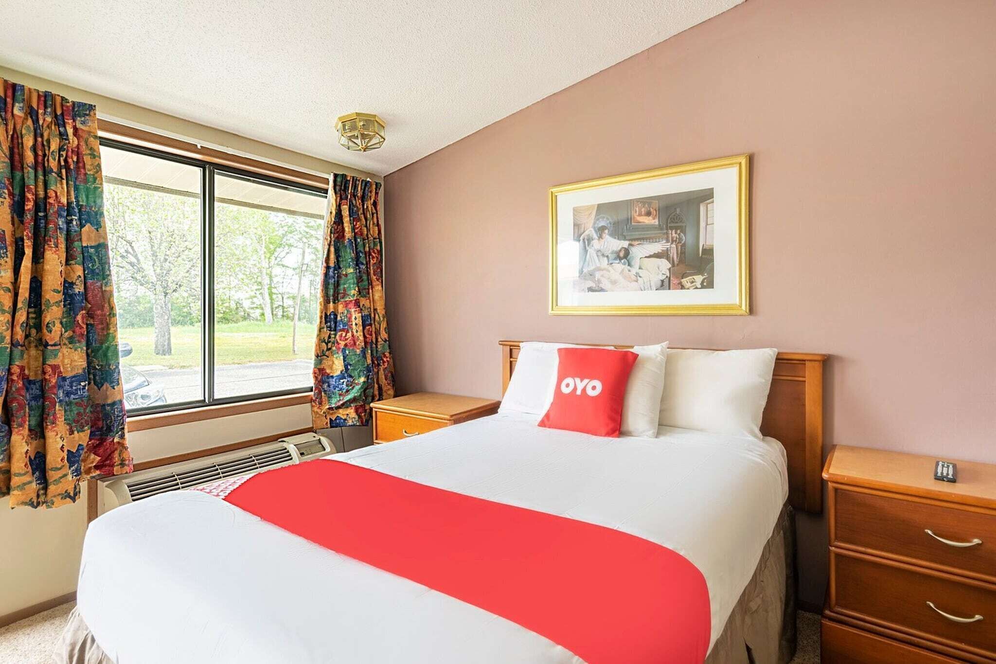 Hotellihuone, jossa on suuri sänky, yöpöytä, seinävalaisin, ikkuna, jossa on verhot, ja seinällä oleva kehystetty kuva.