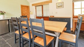 A dining area with a wooden table and chairs, a clock on the wall, and a window with blinds. - Little Trethellan (Redruth)