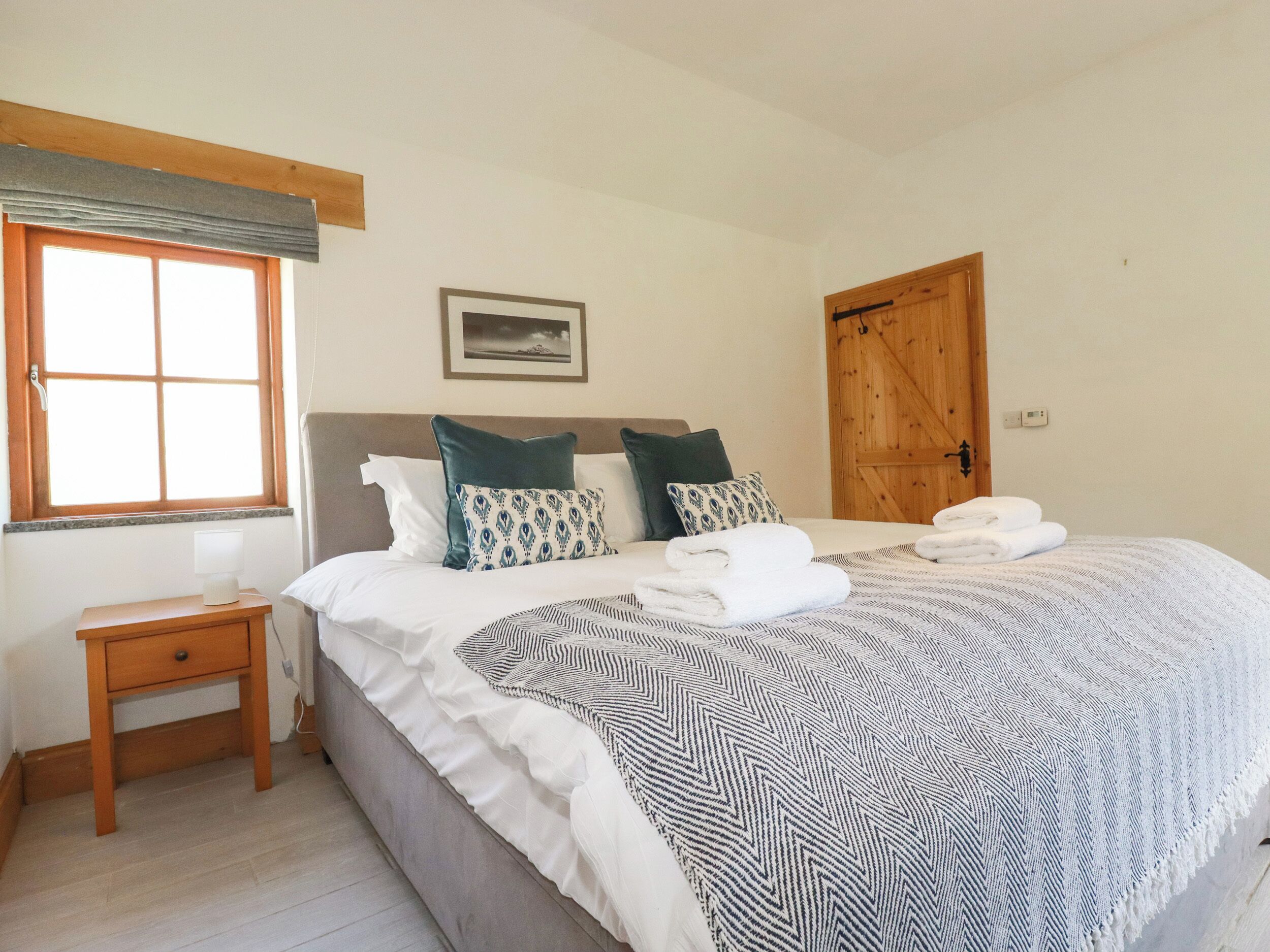 A bedroom with a bed, a bedside table, a window, a door, and a wall-mounted picture.