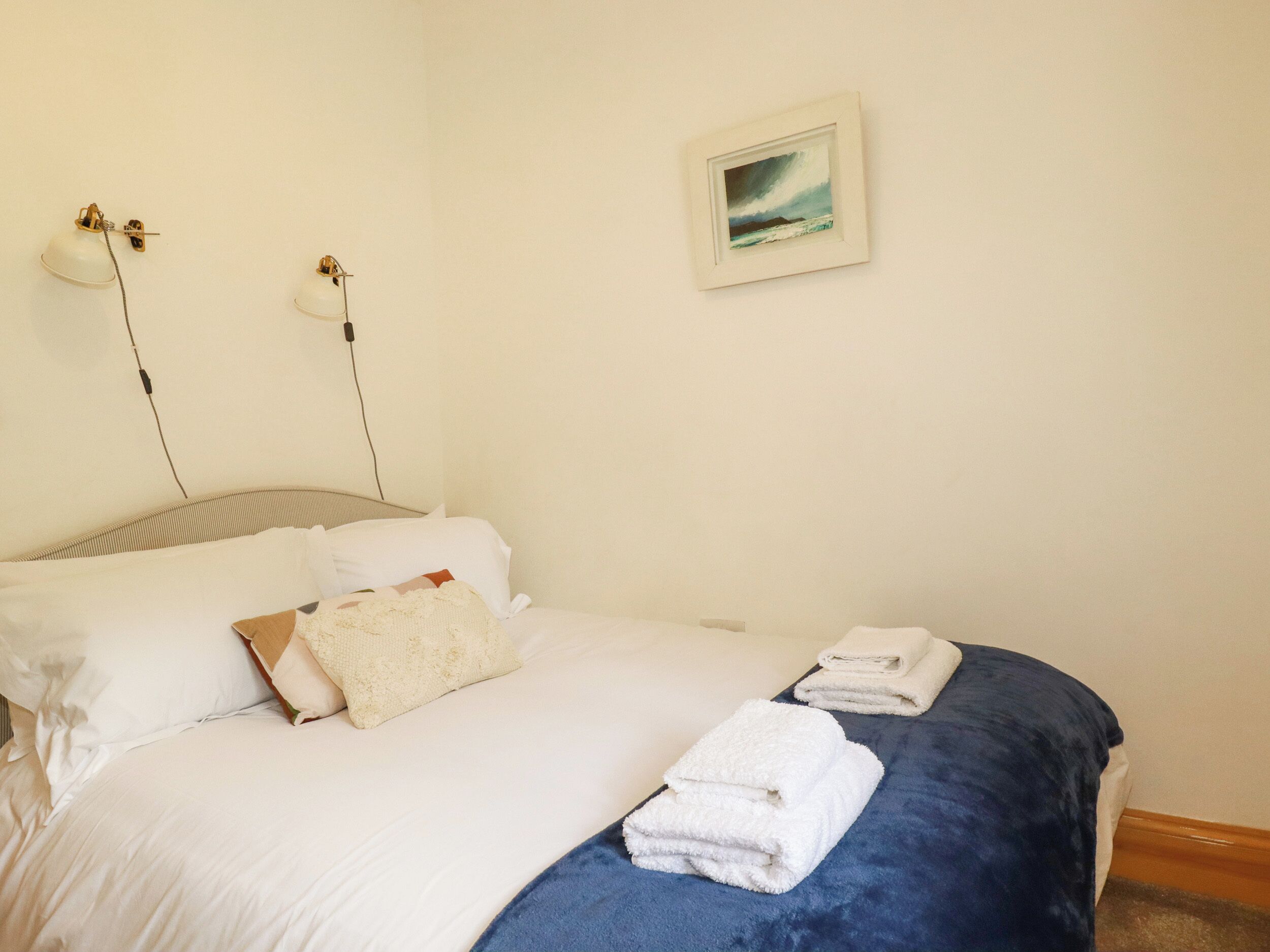 A neatly made bed with white linens, a blue blanket, and folded towels. There are two wall-mounted lamps on either side of the bed.
