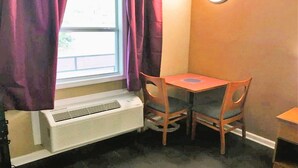 A room with a window, purple curtains, a table with two chairs, a wall-mounted round mirror, and a white air conditioning unit.