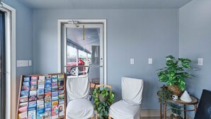 A room with two white chairs, a small table with a plant, a bookshelf with travel books, and a view of a red car through a doorway. - Premium 2 King Bed with Kitchenette (Rockport)