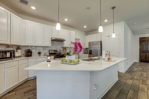 Uma cozinha moderna com armários brancos, uma ilha grande, eletrodomésticos de aço inoxidável e iluminação pendente.
