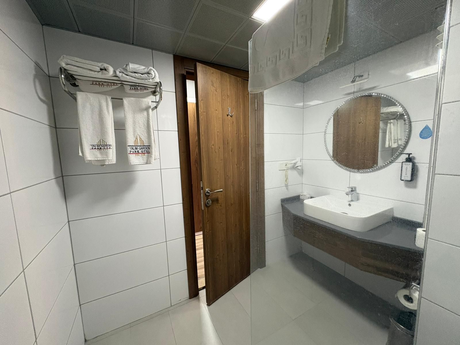 A bathroom with white tiled walls, a wooden door, a sink with a mirror, and towels hanging on a rack.